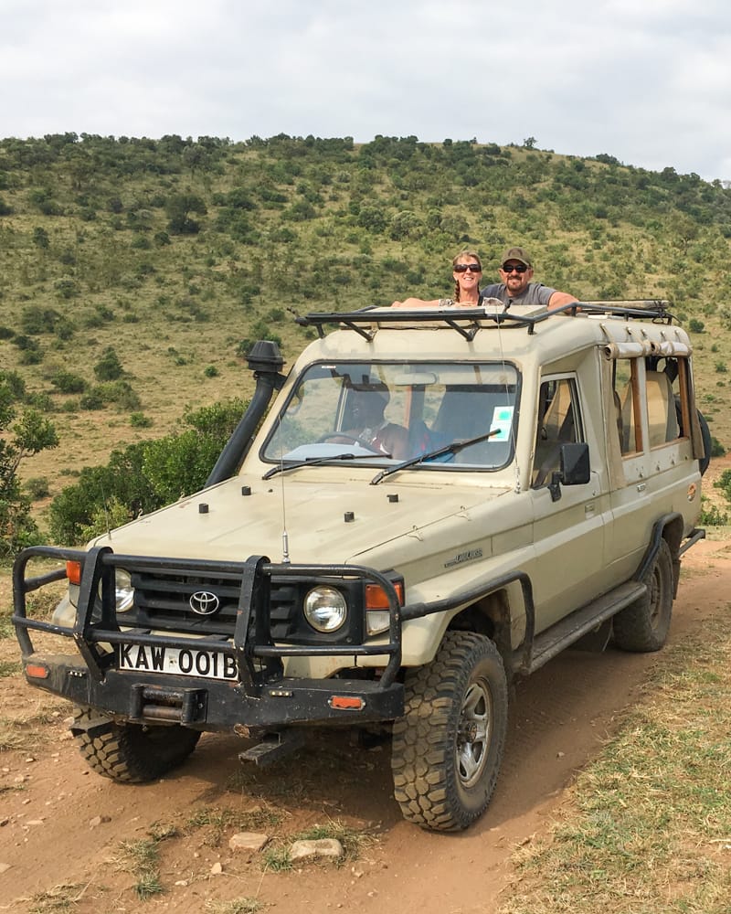 m&e safari vehicle 20 Elaine and Martin standing in the open roof of a beige Toyota Land Cruiser on a dirt track in the Kenyan hills, showing their private safari vehicle during the trip.